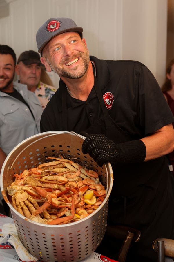 A smiling man in a black shirt and cap holds a large metal basket filled with cooked crab legs and lemons, surrounded by people in a kitchen setting.